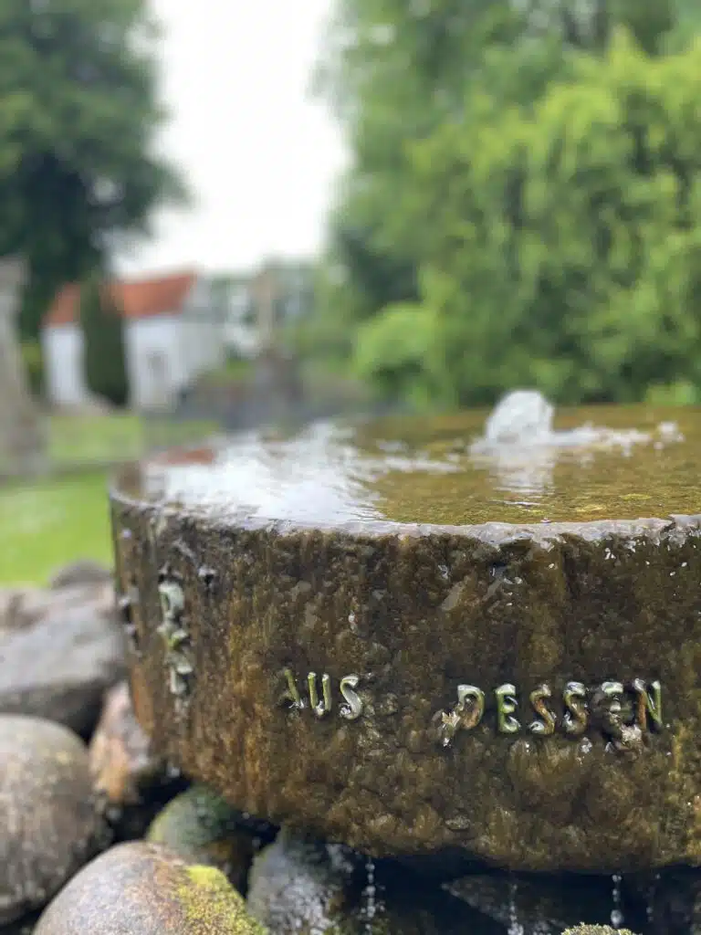 Alter Brunnen mit eingravierter Inschrift auf dem Kirchhof der St.-Johannis-Kirche Adelby, im Hintergrund die weiße Kirche
